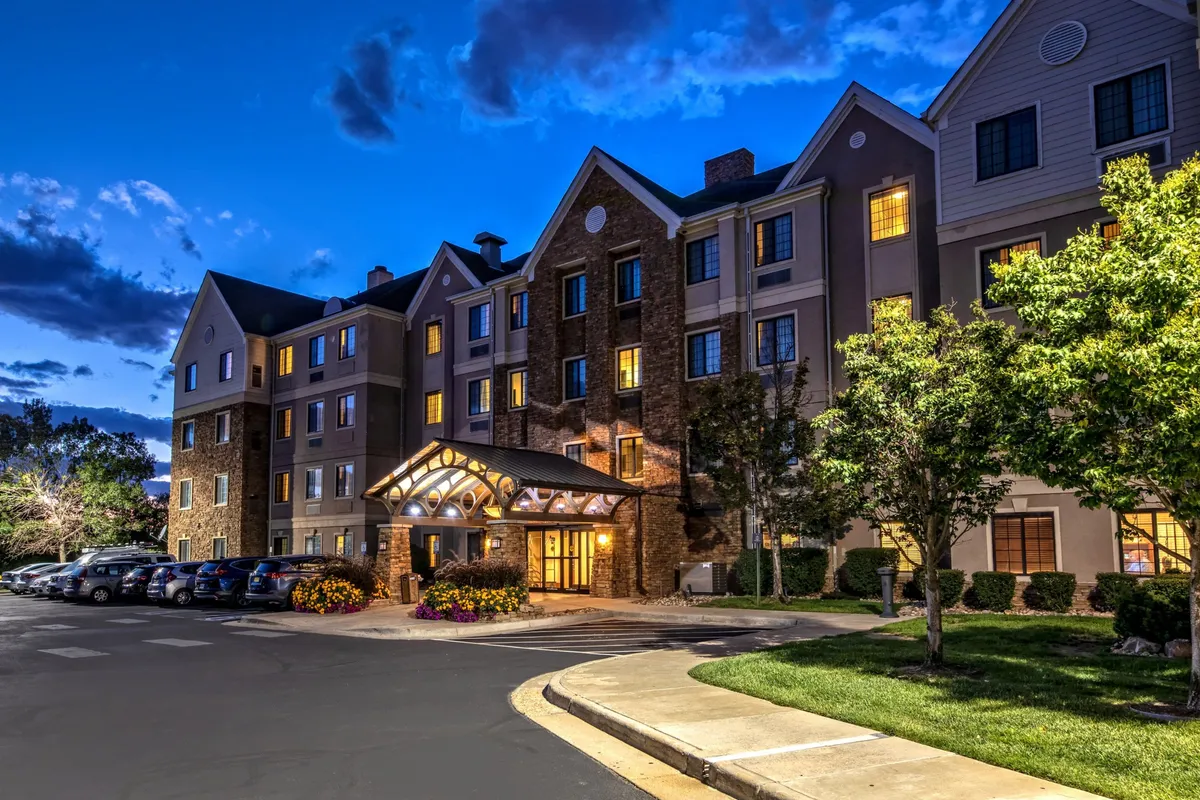 The image features the hotel's entrance awning, warmly lit windows, and a landscaped area in front. The inviting evening ambiance suggests guests returning from nearby attractions and enjoying the welcoming environment.