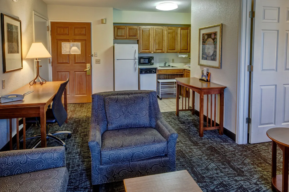 A spacious, sunlit living area featuring a comfortable sofa set, a kitchen area in the background, a dedicated desk with chair for remote work, and colorful artwork on the wall. The open-plan layout invites relaxation, conversation, and productivity, with natural light enhancing the cozy atmosphere.