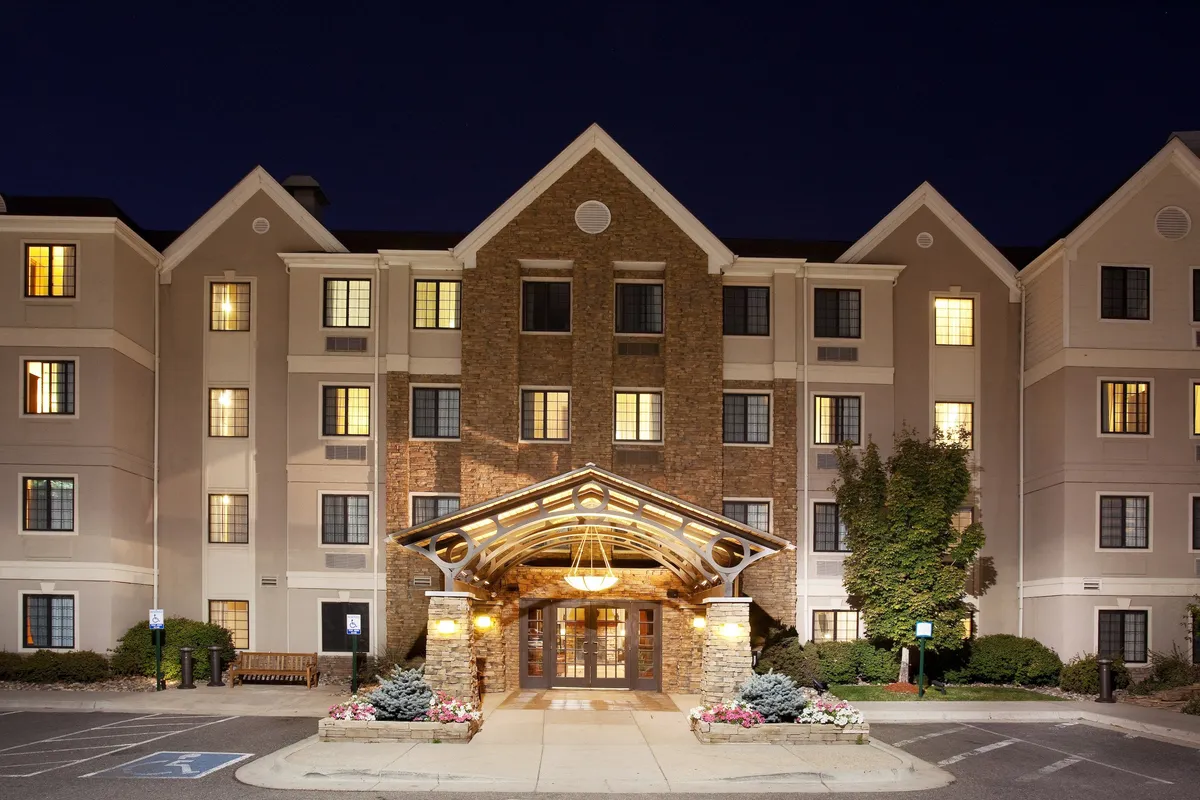 An inviting exterior view of Staybridge Suites Denver-Cherry Creek featuring the well-lit entrance awning, warmly glowing windows, and a landscaped garden setting. The image conveys the welcoming, approachable atmosphere that greets business travelers upon arrival.