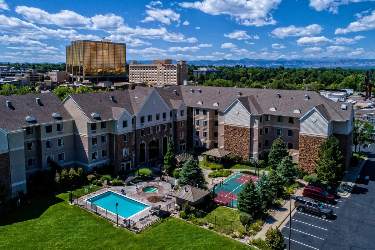 A view of the outdoor swimming pool and adjacent hot tub, surrounded by landscaped gardens and partially visible tennis courts. Guests can be seen relaxing by the pool and enjoying the vibrant, sunny atmosphere, suggesting a lively yet relaxing communal setting.