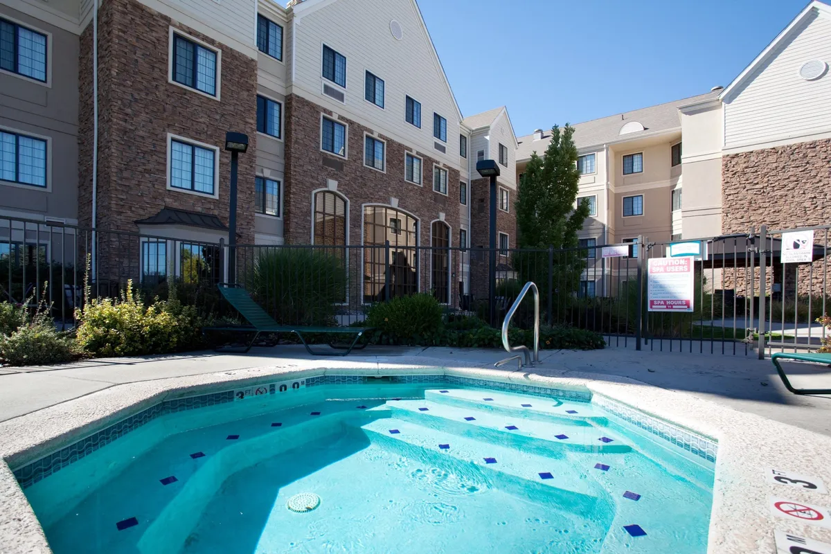 This exterior image showcases the Staybridge Suites Denver-Cherry Creek building with a focus on the inviting hot tub and pool area, surrounded by well-maintained landscaping. Guests can be seen relaxing by the pool, enjoying the outdoors as the sun sets, creating a casual and relaxing mood.