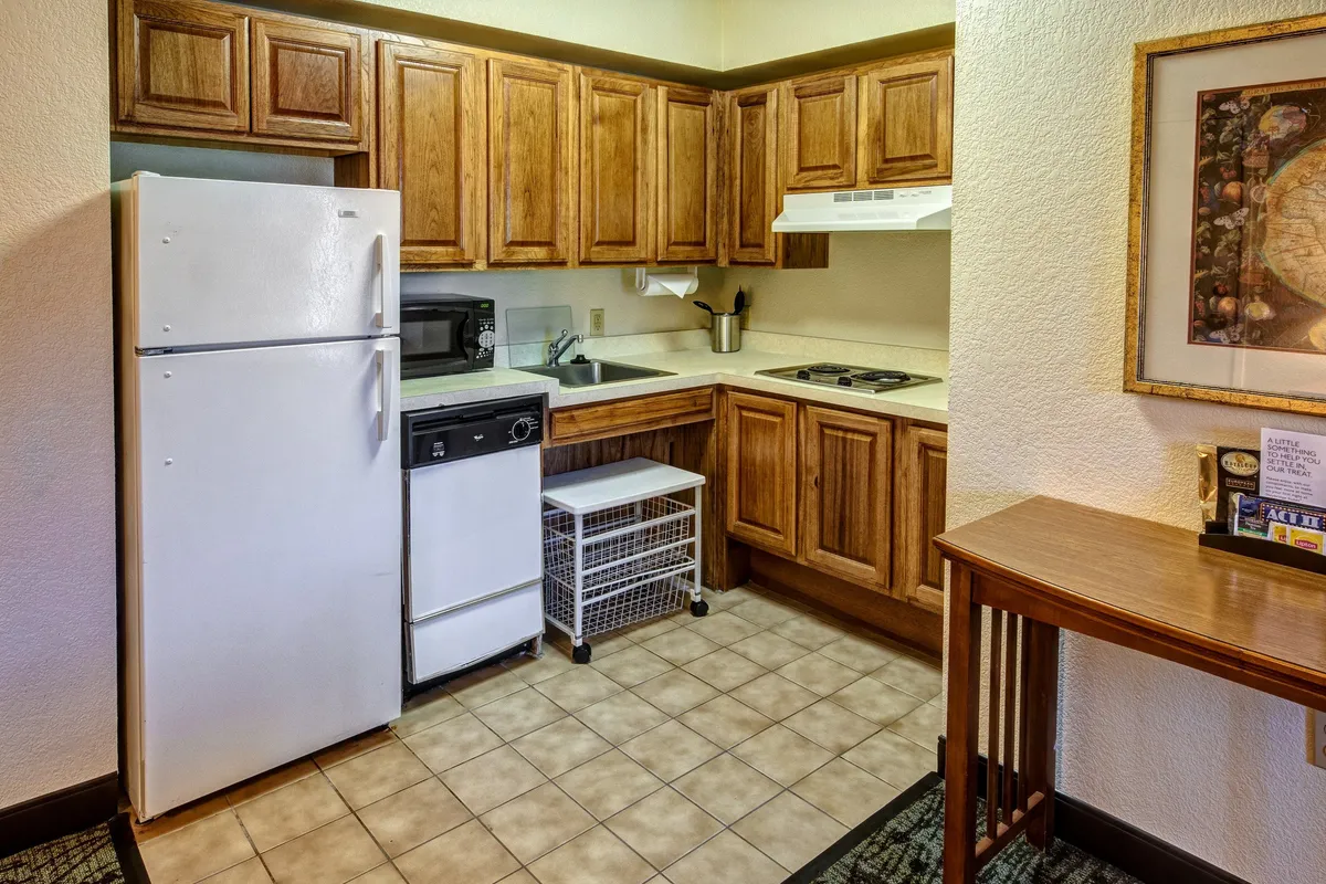 A warm and functional kitchen space featuring a refrigerator, stove, kitchen cabinets, and sink, with a small table nearby. The area is arranged for meal preparation and social gathering, and includes visible cooking utensils and appliances, suggesting readiness for both cooking and dining.