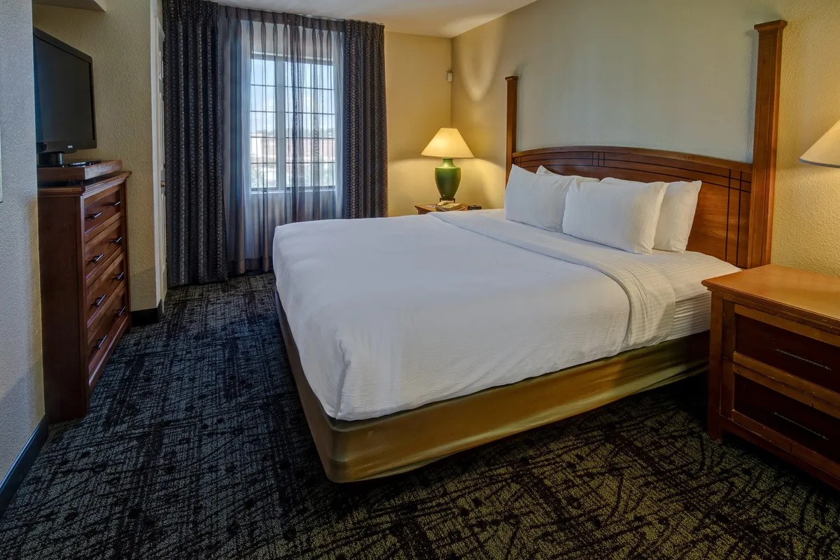 A comfortable bedroom is pictured, featuring a queen-size bed with plush bedding, flanked by nightstands and a lamp. A television is mounted for private entertainment, and the room is brightened by natural light streaming through a window with soft curtains.