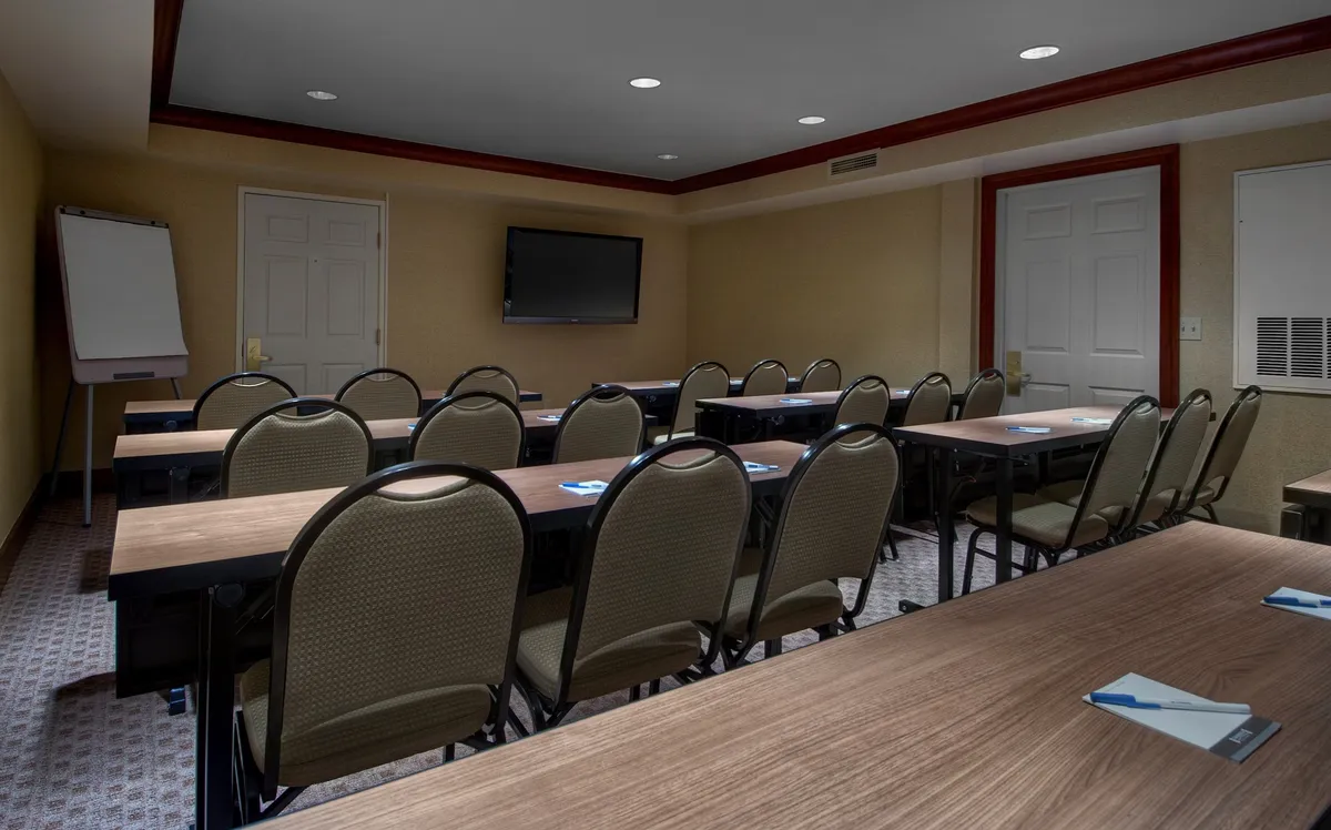The image depicts a well-lit, modern meeting room with rows of tables and chairs, facing a television screen mounted on the wall and a flip chart easel. The setup is ideal for collaborative activities, presentations, and group gatherings, fostering a productive yet comfortable atmosphere.